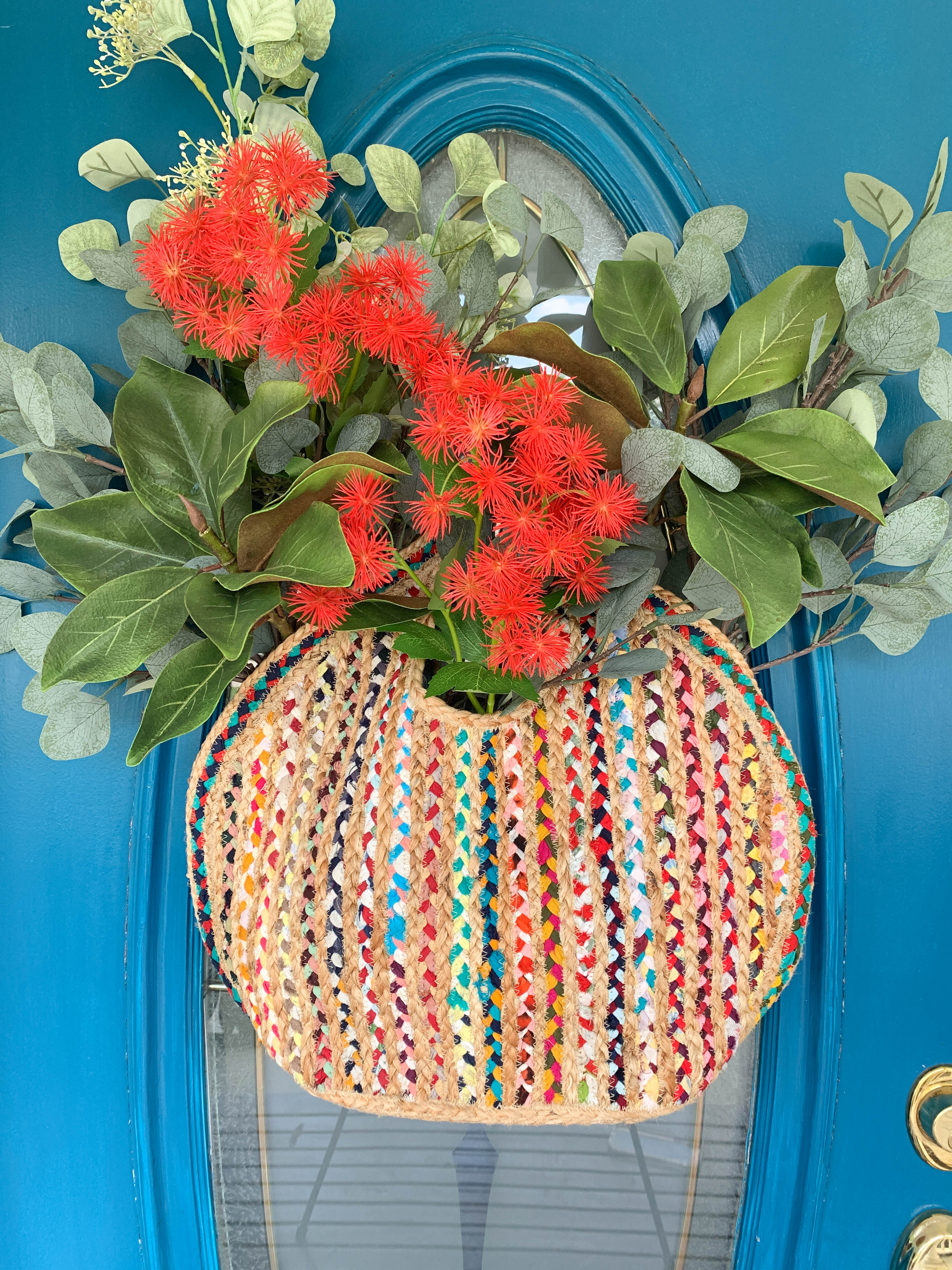A vibrant orange-red flower tucked in amidst faux greenery in a natural weave handbag hanging on an entrance door.