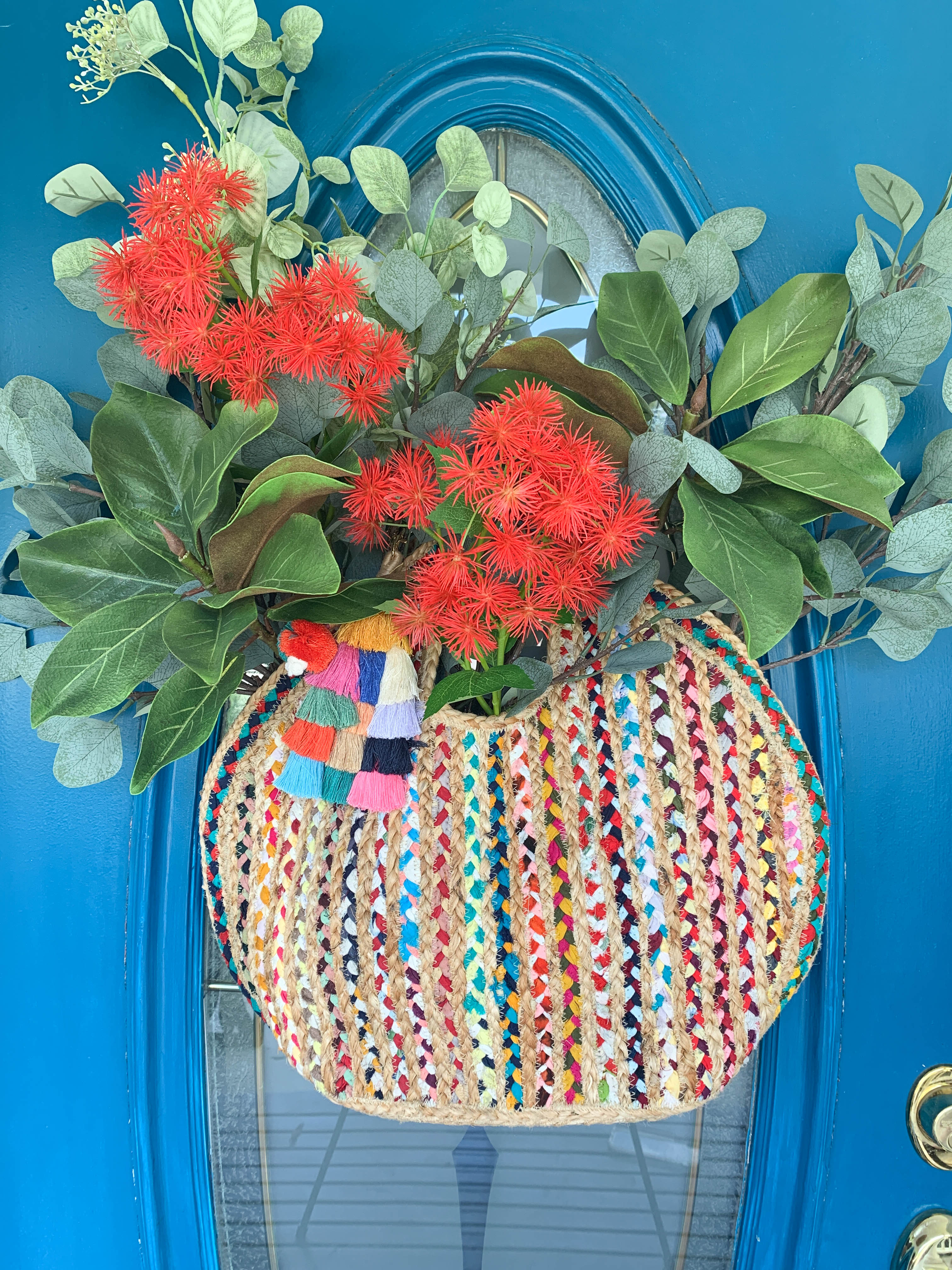 A multi-colored woven bag full of faux greenery and flowers and hanging on the front door as decor.