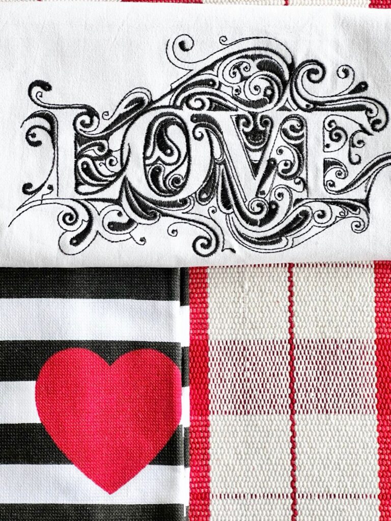 Layers of linens including an embroidered "Love" tea towel. a heart tea towel and a red gingham table runner.