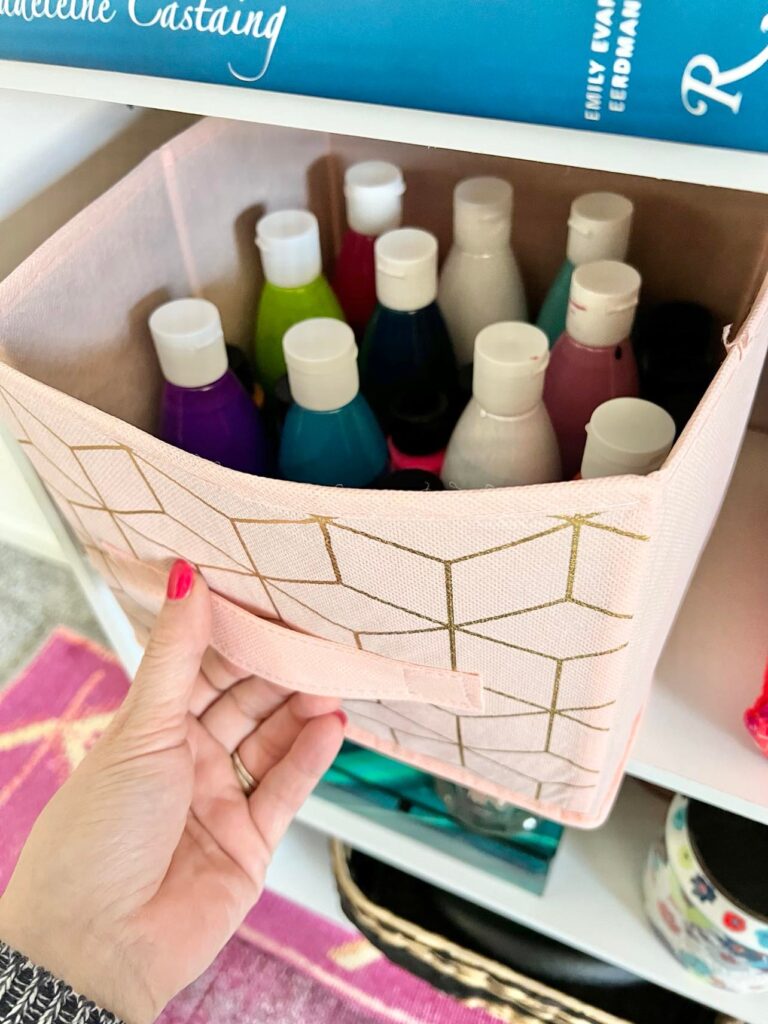 A pink fabric bin holding bottles of craft paint.