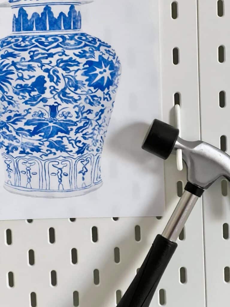 A hammer and blue and white art print hanging on a pegboard.