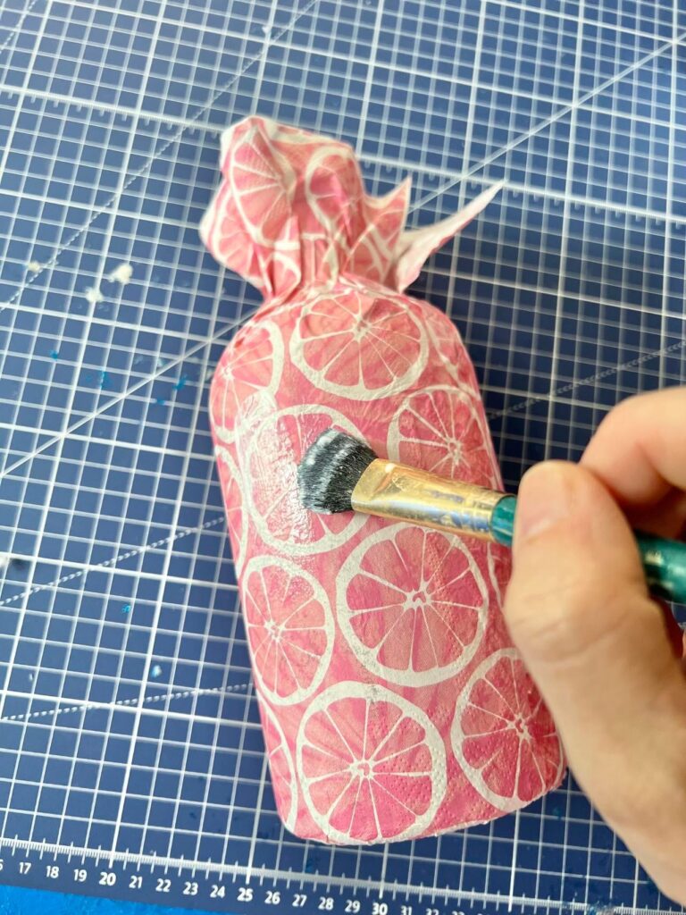 A brush applying a thin coat of glue over the bottle covered in a paper napkin.
Decoupage bottle idea.