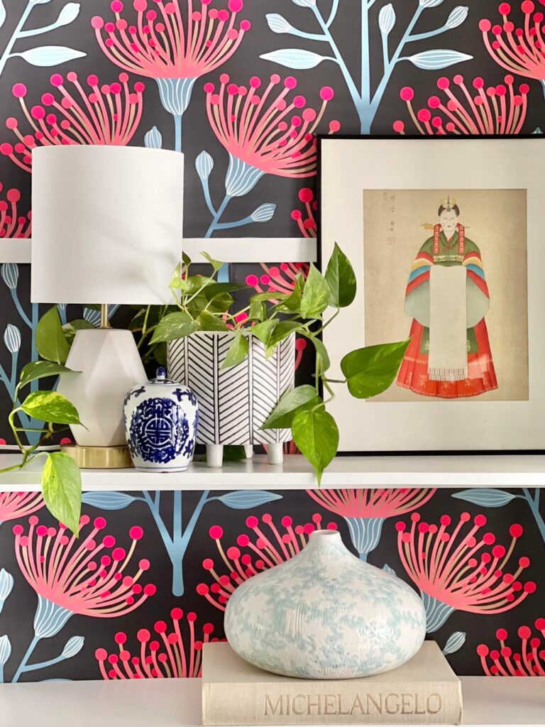 Floral wallpaper lines the back of a bookcase that is styled with colorful home decor that includes a lamp, plant, and artwork.