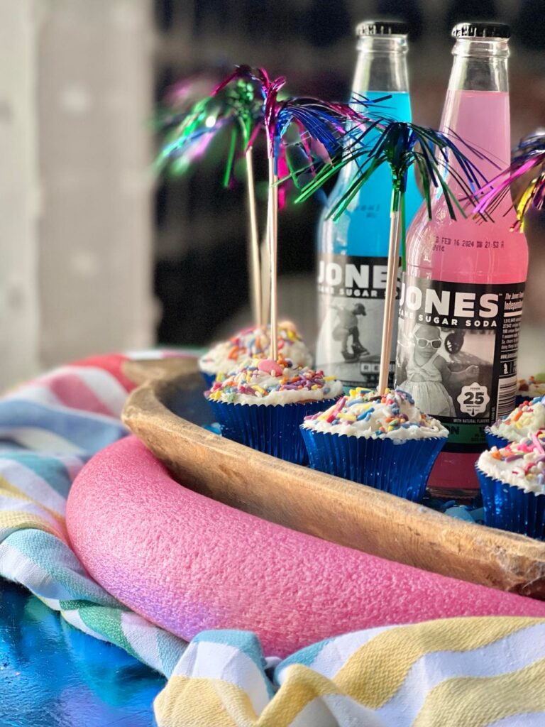 A dough bowl filled with glass color pebbles, with cupcakes and sodas sitting on top.