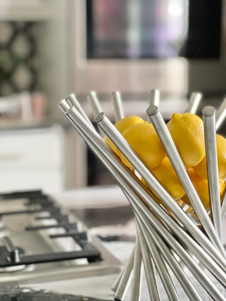 An open weave stainless steel fruit bowl holding yellow lemons and sitting on top of the kitchen stovetop.