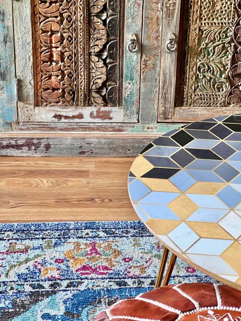 A carved chest sitting on a wood floor with a round table and leather ottoman sitting on a rug in the colors of navy, gold, and fuschia.