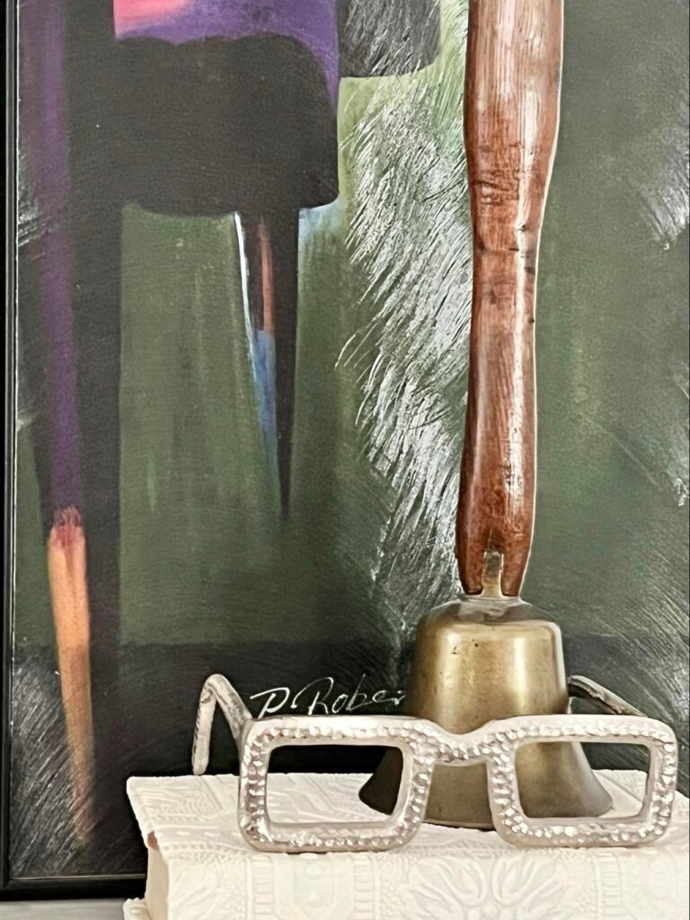 An antique school teacher's bell sitting on a stack of books with some home decor eyeglasses.