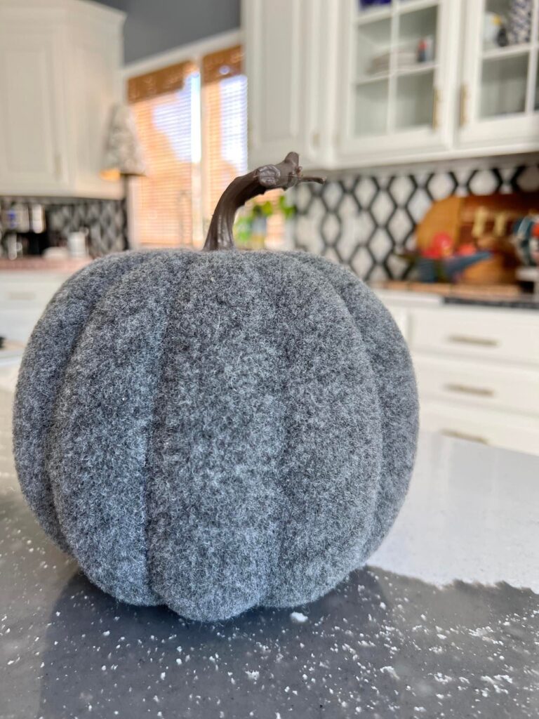 A grey fuzzy faux pumpkin with a plastic stem.