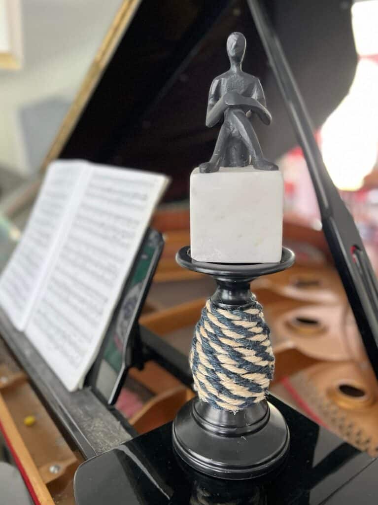 The jute wrapped candlestick sitting on a piano with a black and white sculpture sitting on top.
