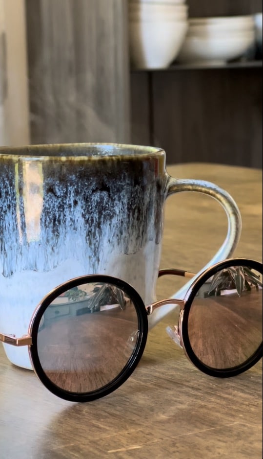 A cup of coffee sitting on kitchen table beside a pair sunglasses.