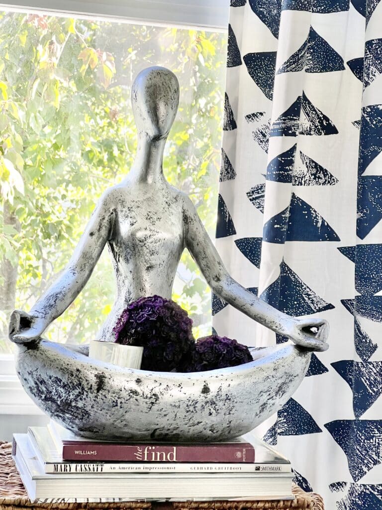 A lotus position sculpture sits in a window with a blue and white curtain backdrop.