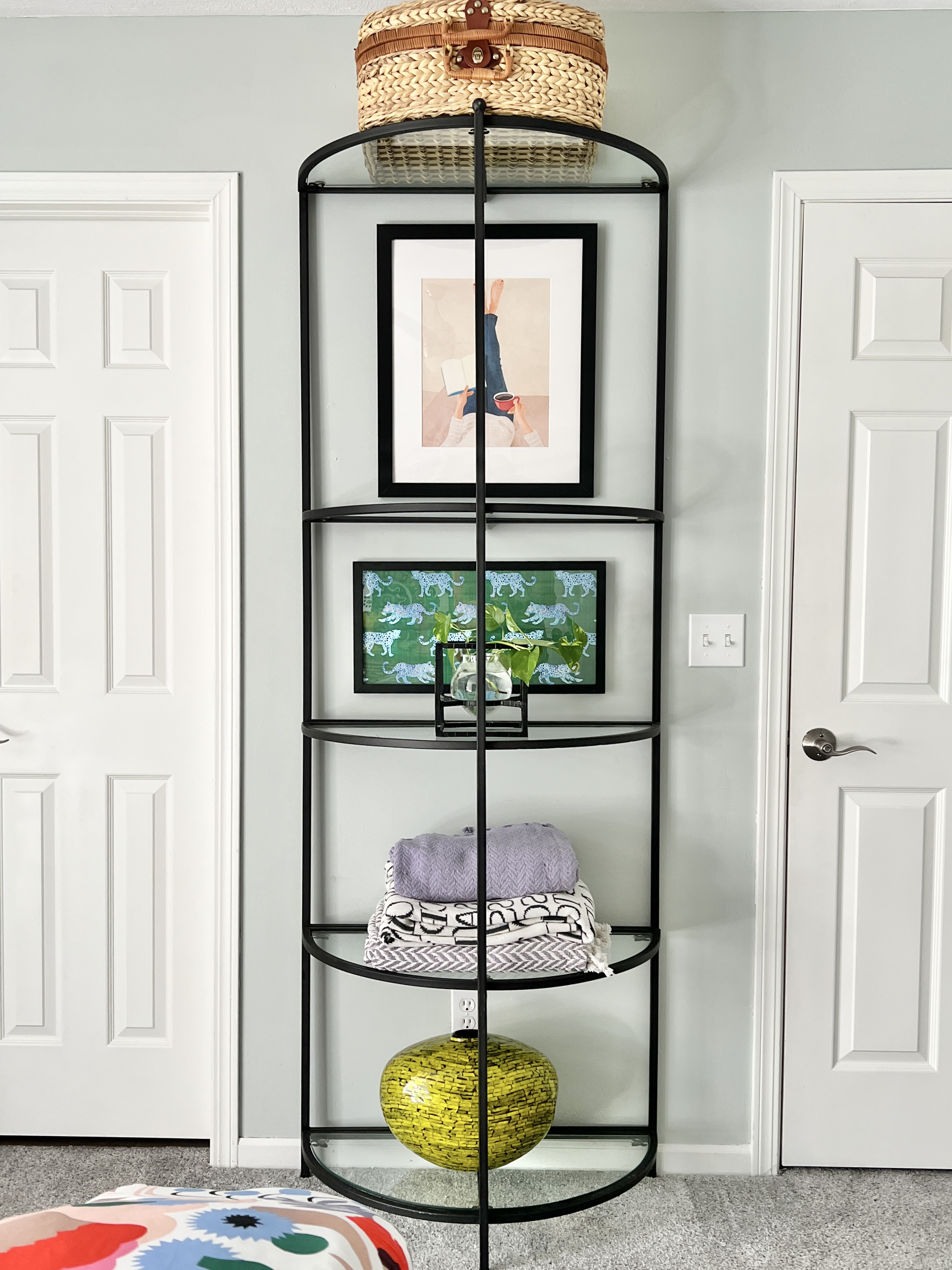 A metal and glass etagere serves as a bookcase to hold more wall art, extra blankets, and give guests storage options in their cozy guest room.