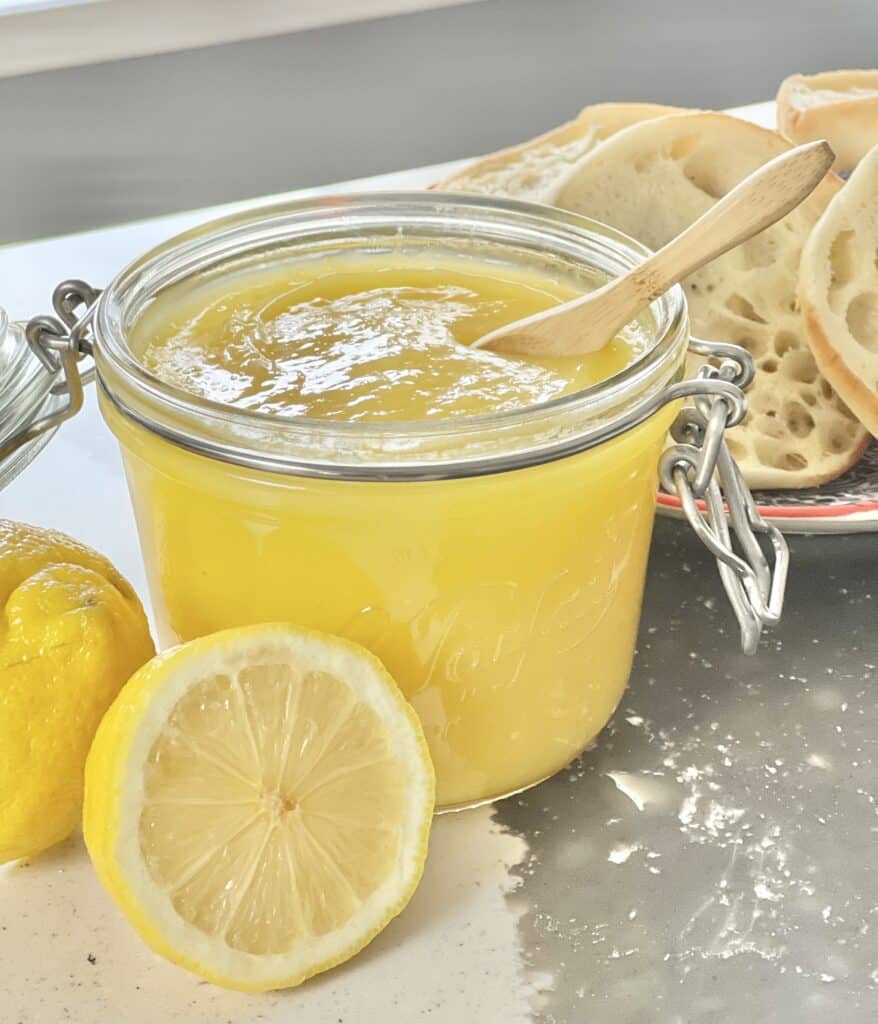 A jar of lemon curd sitting on a counter beside cut lemons and a plate of english muffins.