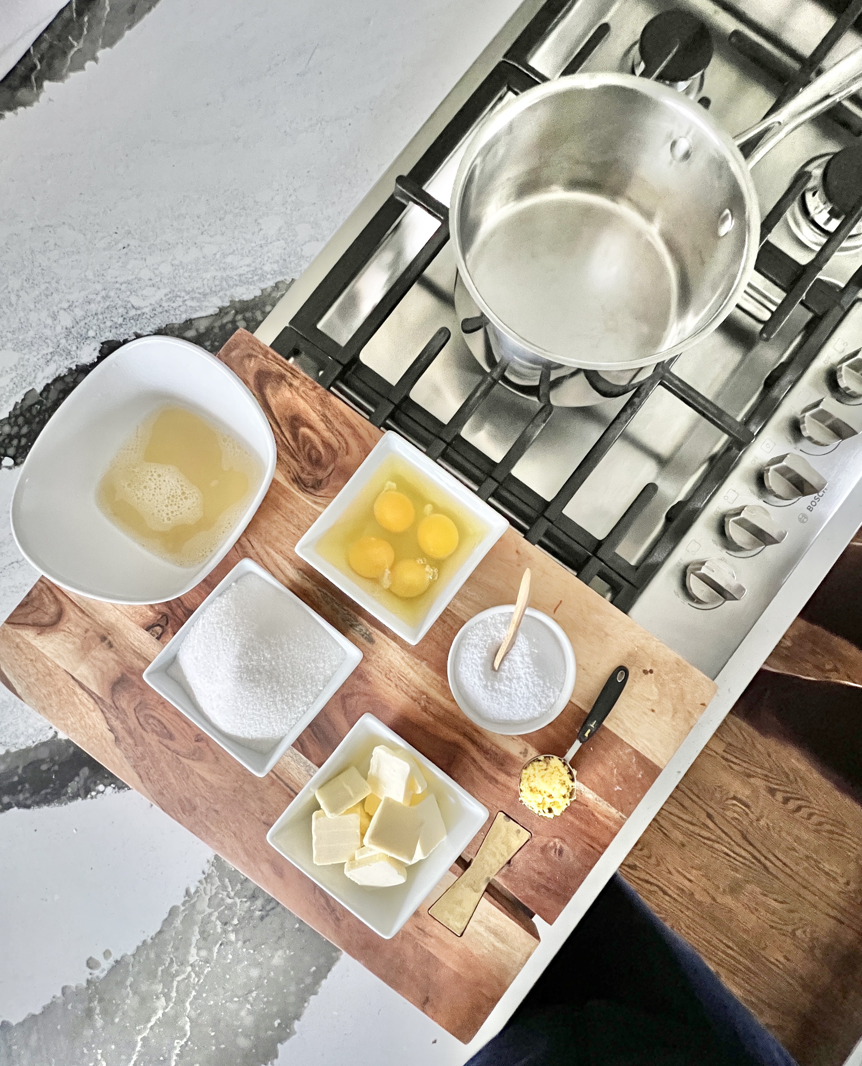 A pan on the stove beside bowls of lemon juice, sugar, eggs, butter, salt, and lemon zest.
