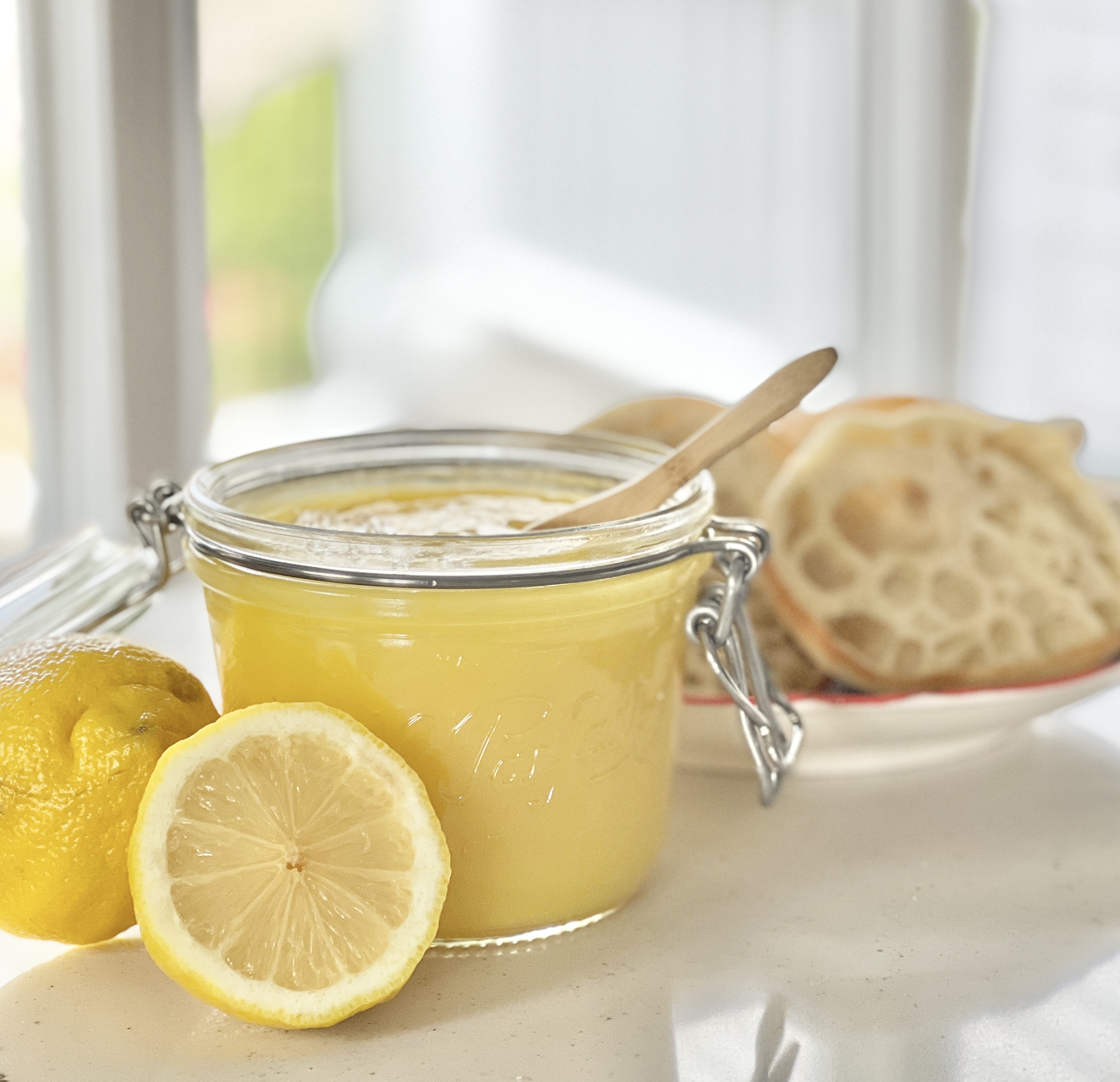 Lemon curd in a glass jar beside a plate of english muffins.