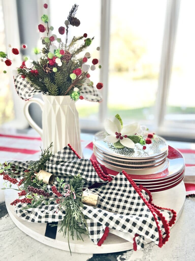 A Christmas centerpiece idea consisting of stacked plates, piles of napkins, and Christmas florals in white pitchers.