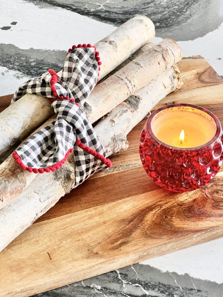 A lighted candle sitting on a wood kitchen cutting board and some birch logs tied with a decorative napkin.