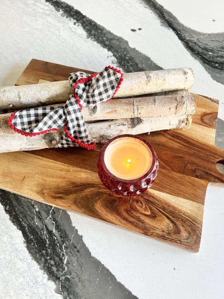 A bunch of birch logs tied with a simple gingham checked napkin with a candle on top of a charcuterie board.