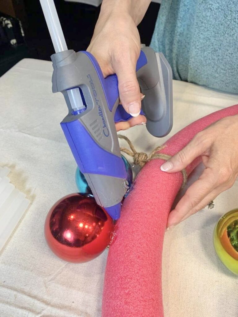 Christmas ornament wreath DIY: Using a hot glue gun to attach ornament balls to the pool noodle wreath form.