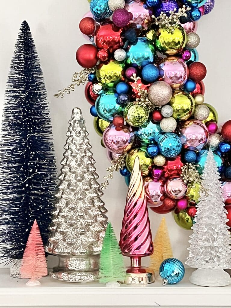 A pool noodle ornament wreath surrounded by glass trees on a fireplace mantel.