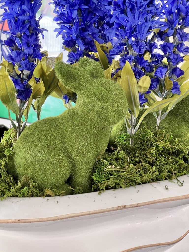 A moss covered bunny sitting in a white planter with faux flowers.