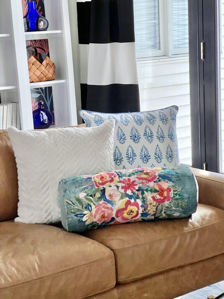 Two colorful throw pillows are combined with a white pillow on a brown leather sofa.