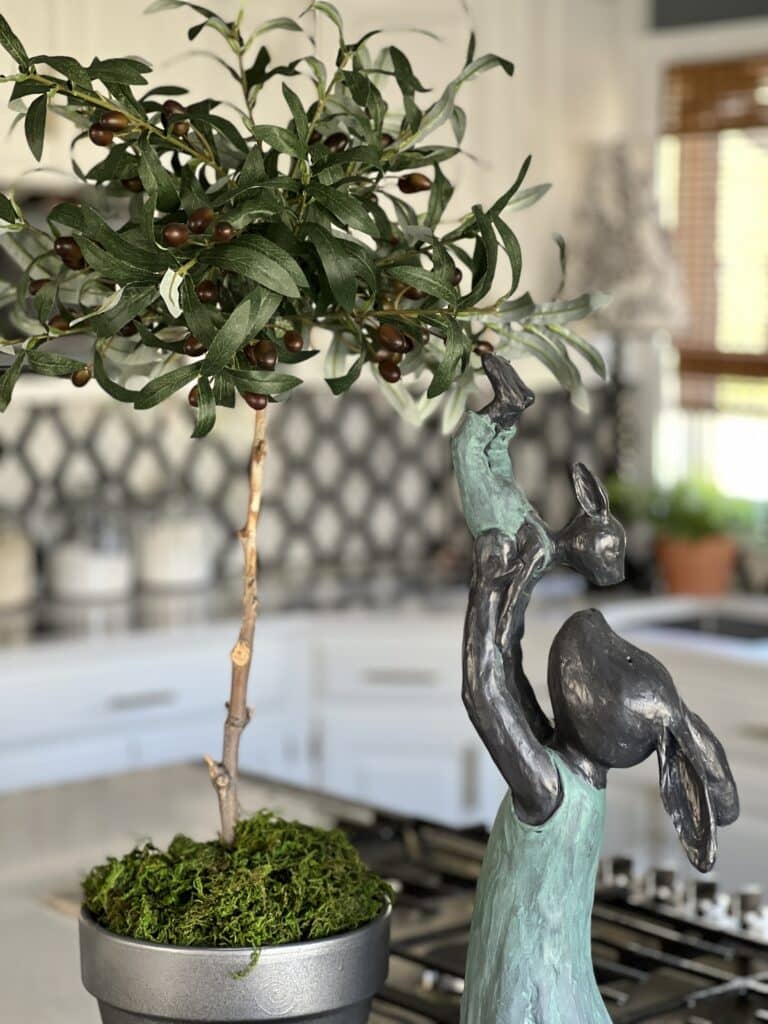 A faux olive tree topiary displayed on a kitchen island with a bunny sculpture.