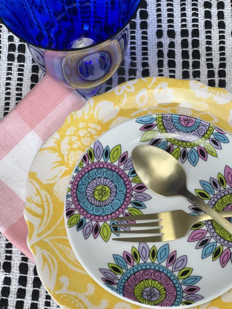 A colorful place setting includes a pink napkin, yellow plate, gold fork and spoon, and pink patterned plate.