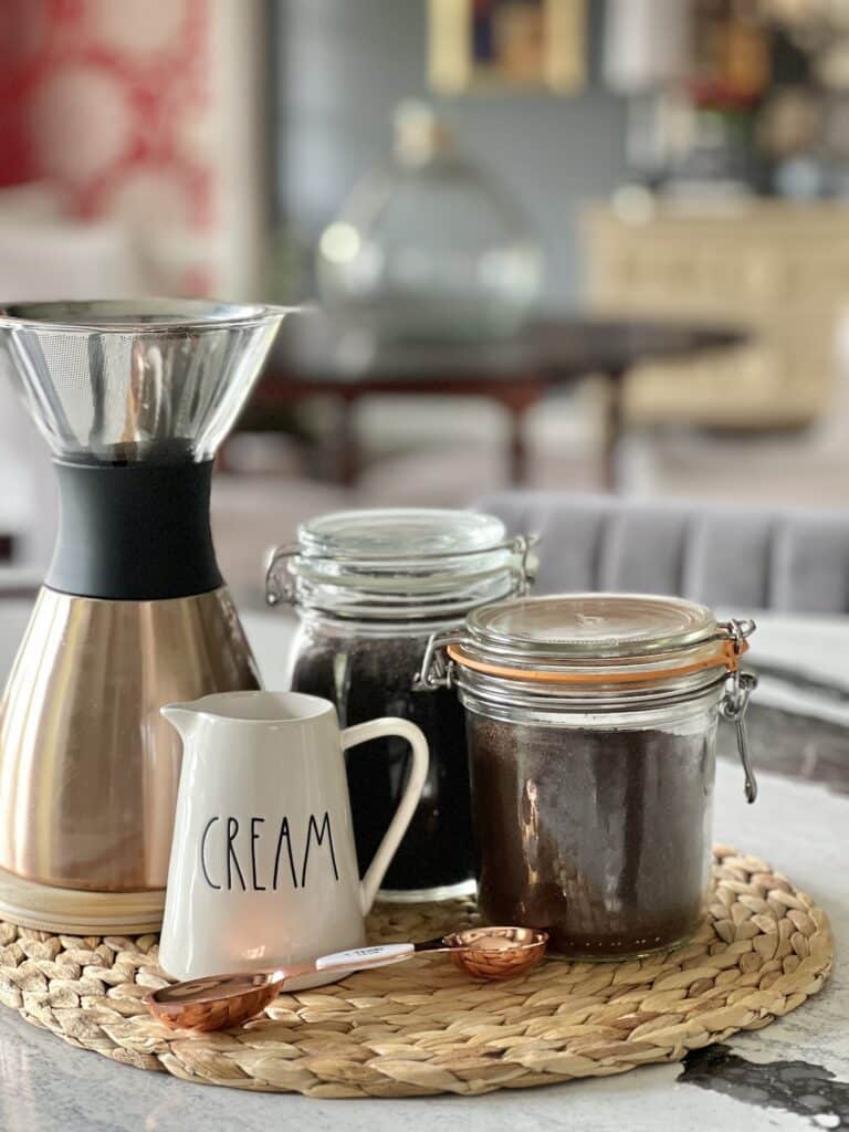Sealed glass container holding ground coffee is displayed on a woven placemat with a coffee pot and cream pitcher.