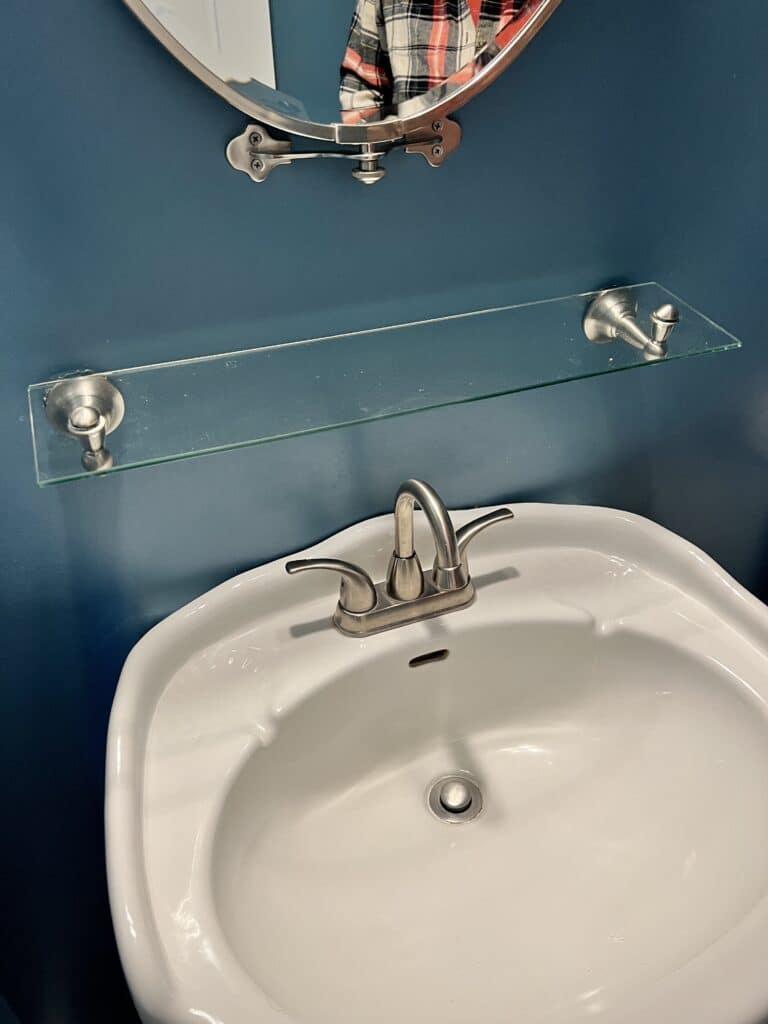 A glass shelf attached to the wall above the half bath sink.