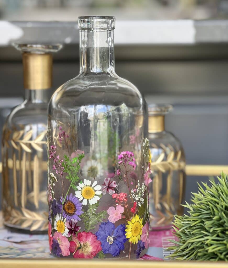 A pressed flower bottle vase displayed among other glass decor in on a kitchen bar cart.