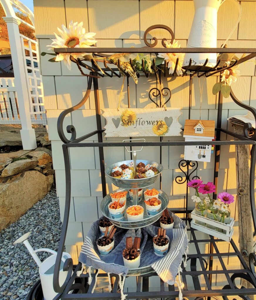 A bakers rack sitting outside and decorated with flowers and sweet treats for entertaining.