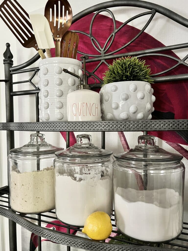 Canisters of flour and sugar sit beside a crock holding utensils in this example of decorating a kitchen bakers rack.