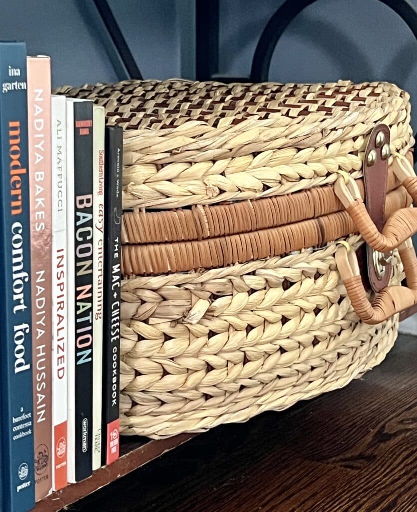 Cookbooks sitting on a shelf beside a wicker woven picnic basket.