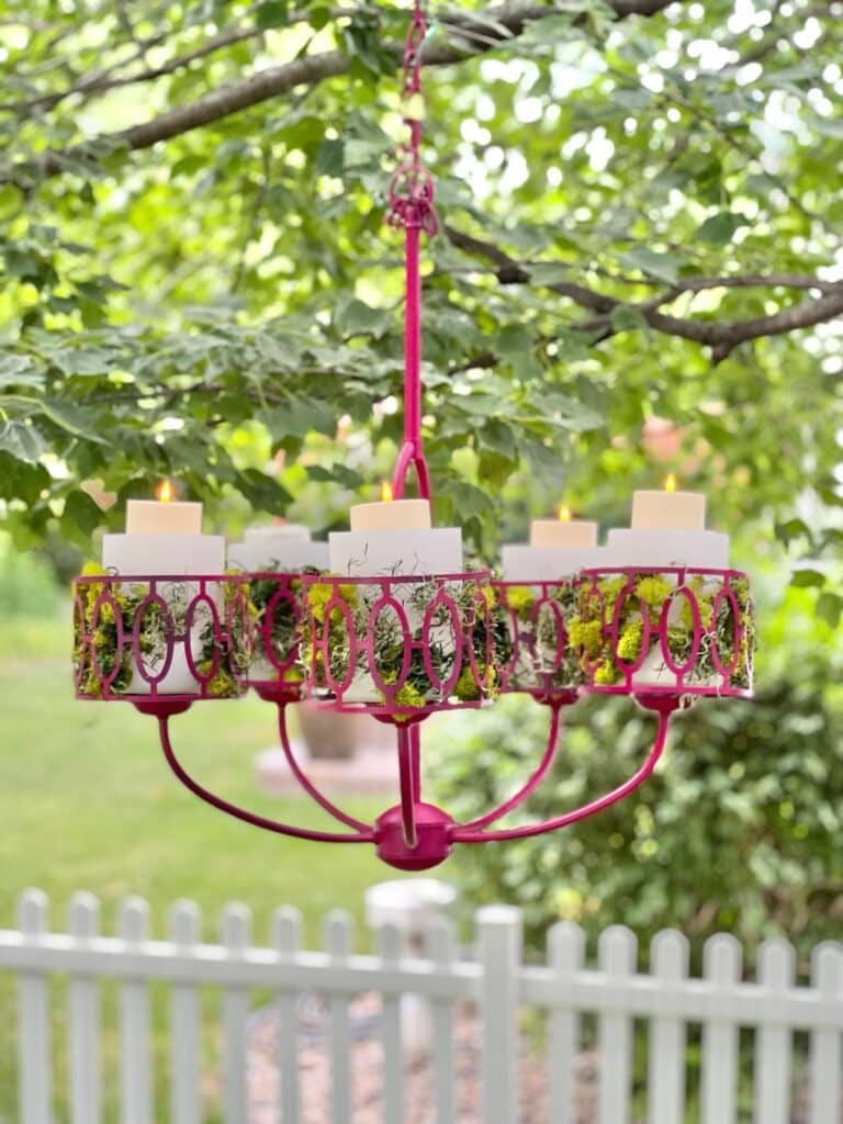 A metal light fixture that has been painted a magenta color and repurposed as an outdoor chandelier hanging from a tree branch.