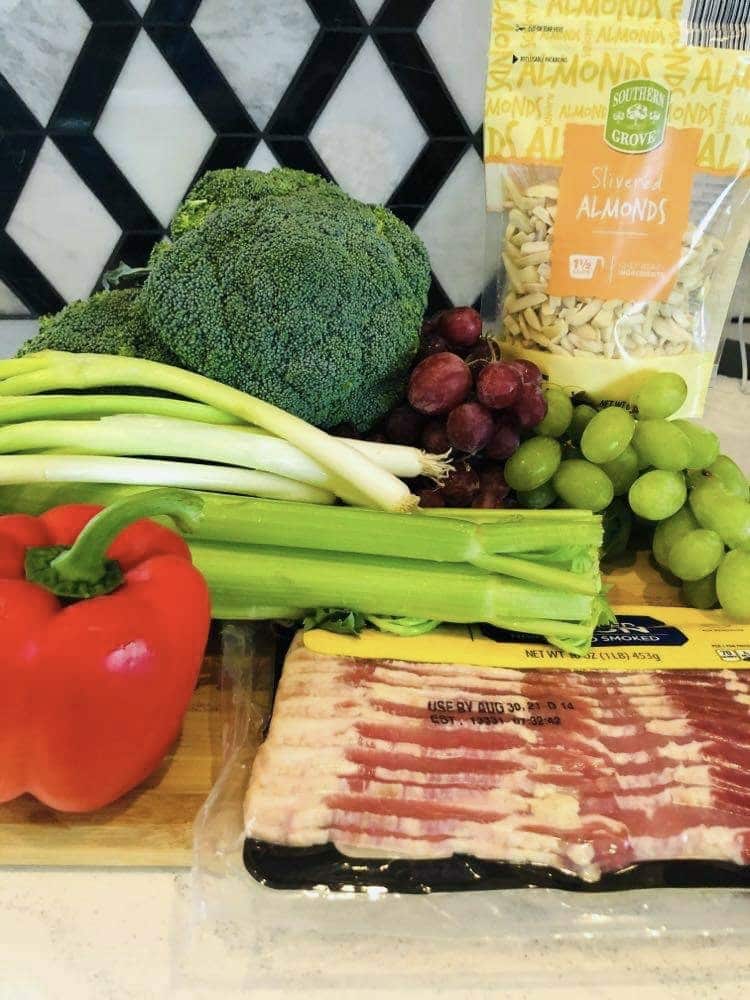 The ingredients for the broccoli grape salad that includes red bell peppers, broccoli, green onion, re and green grapes, bacon, and almonds.