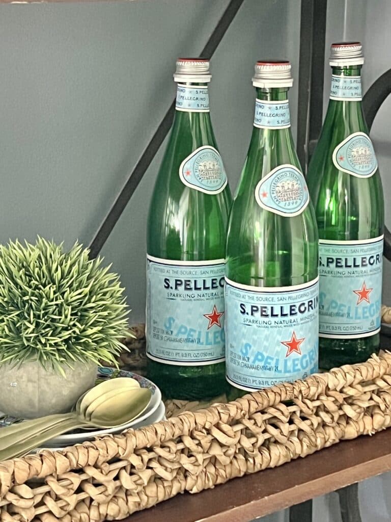 Three bottles of sparkling water tucked in a woven basket beside a faux plant.