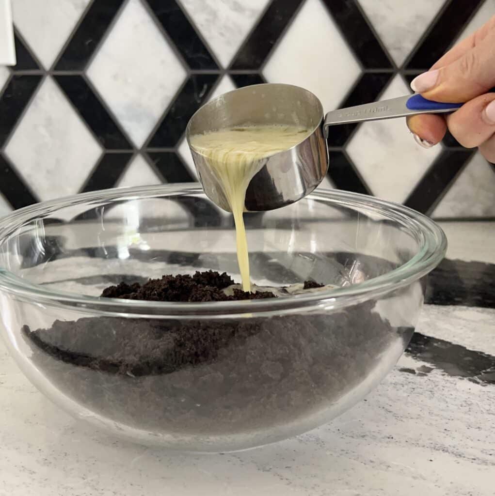 Pouring melted butter onto crushed chocolate cookies for the Buster Bar recipe crust.