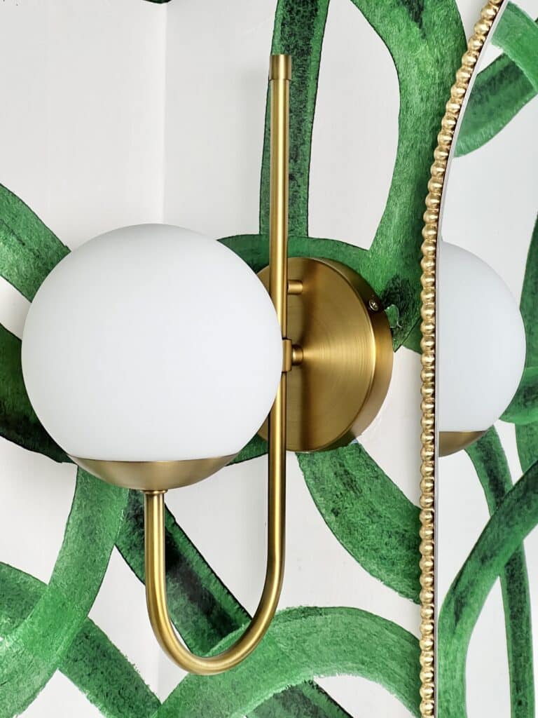 how high to hang bathroom sconces: A wall sconce with gold curving arms and a white globe hanging on the wall beside a mirror.