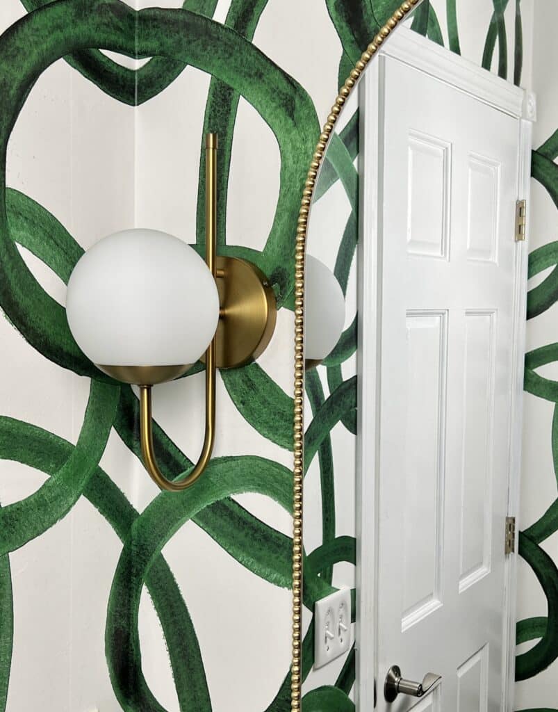 A bathroom wall sconce on the wall beside a gold beaded mirror.