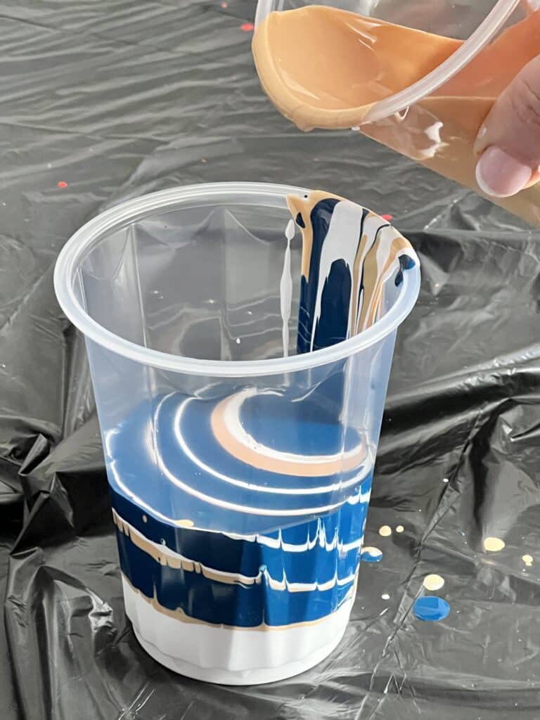 DIY abstract wall art: Pouring layers of white, blue, and gold into a plastic cup in preparation for a paint pour on the canvas.