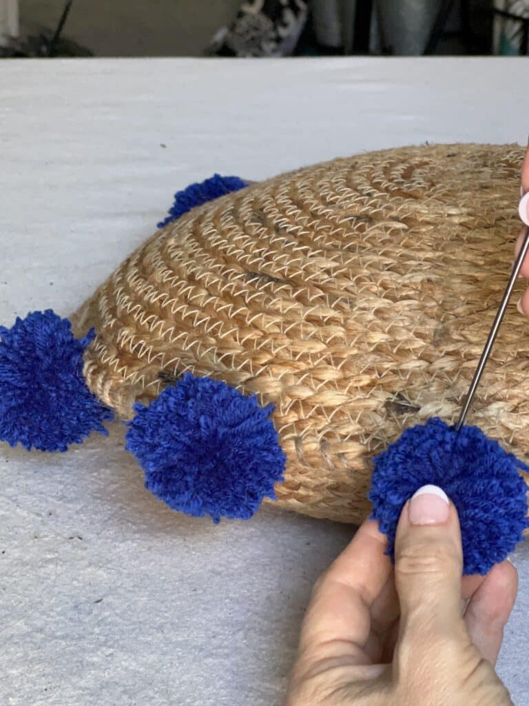 Using the upholstery needle to attach pom poms to the perimeter of the DIY boho pillow.