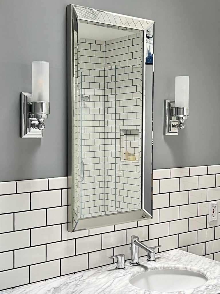 how high to hang bathroom sconces: Two wall sconces with a chrome finish on either side of a glass framed mirror.