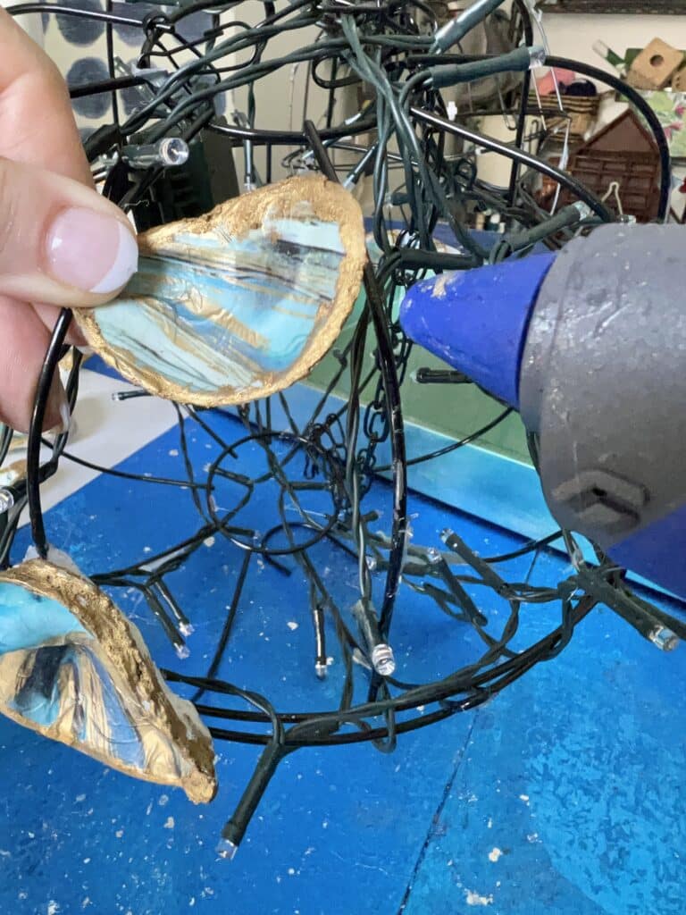 Using hot glue to attach oyster shells to a wire basket globe to create a DIY light chandelier.