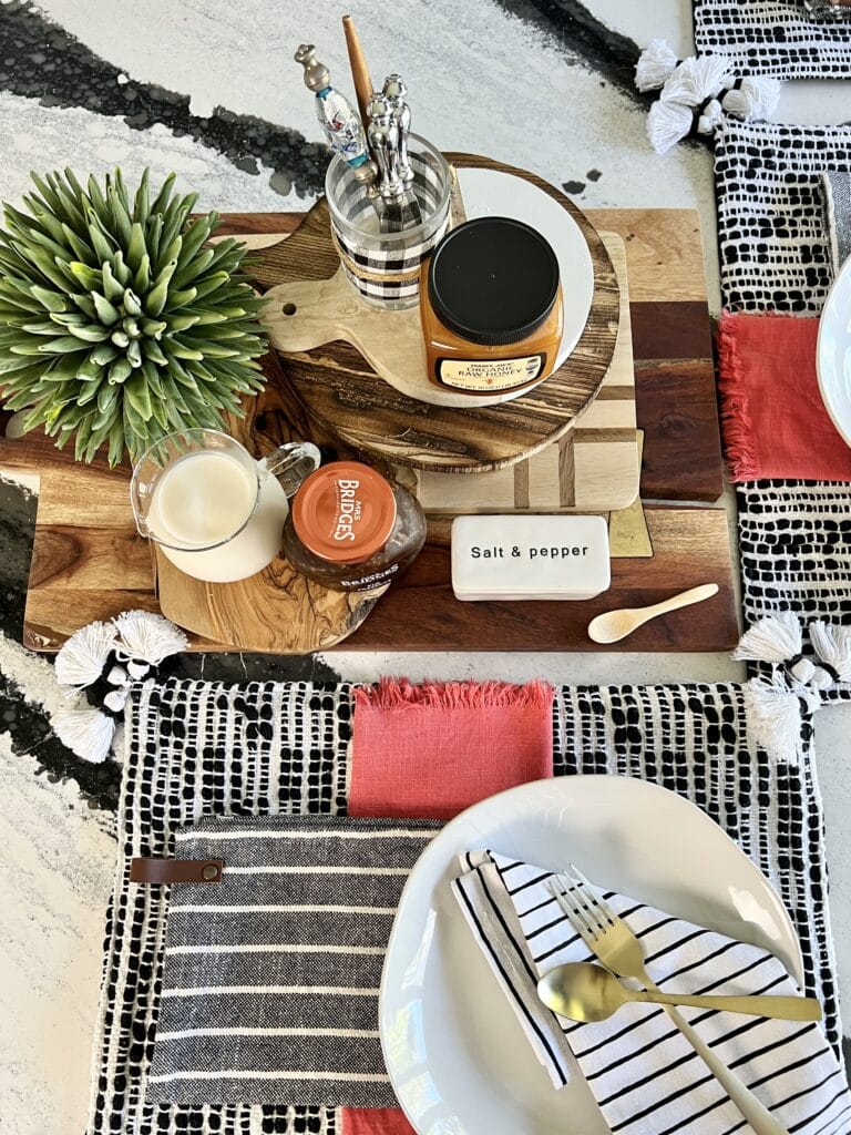 Several wood cutting board stacked vertically and used as a centerpiece to hold a pitcher of milk, jam jars, and salt and pepper.