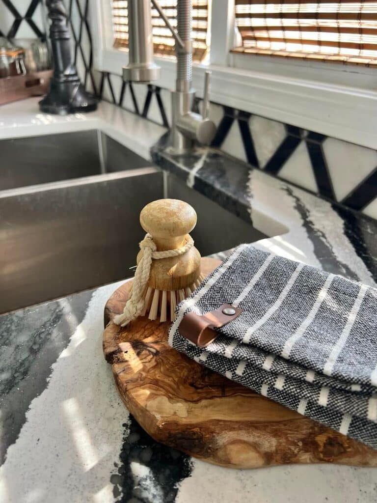 A grey and white striped tea towel folded on top of a wood cutting board and displayed with a wood handled brush.