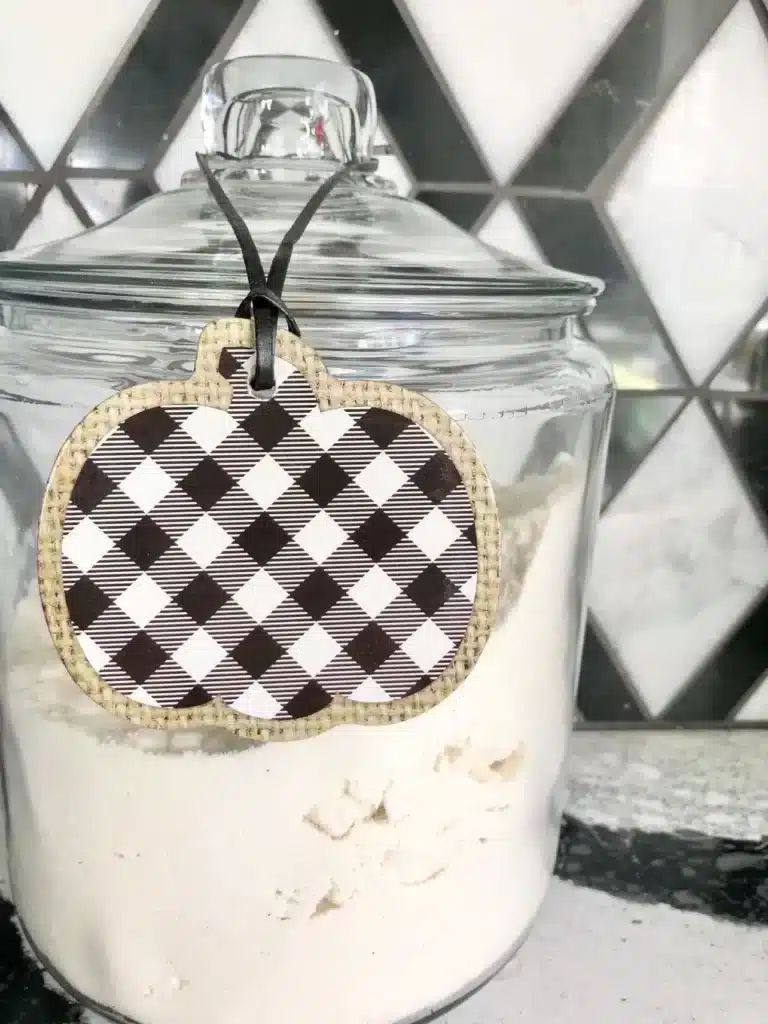 kitchen fall decor ideas: A black and white gingham pumpkin tab hanging from the lid of a clear glass flour canister.