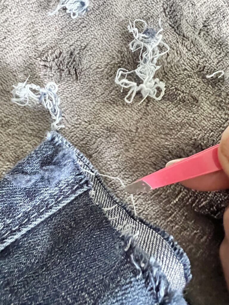how to fray jeans: Using tweezers to pull individual threads from the denim hem.