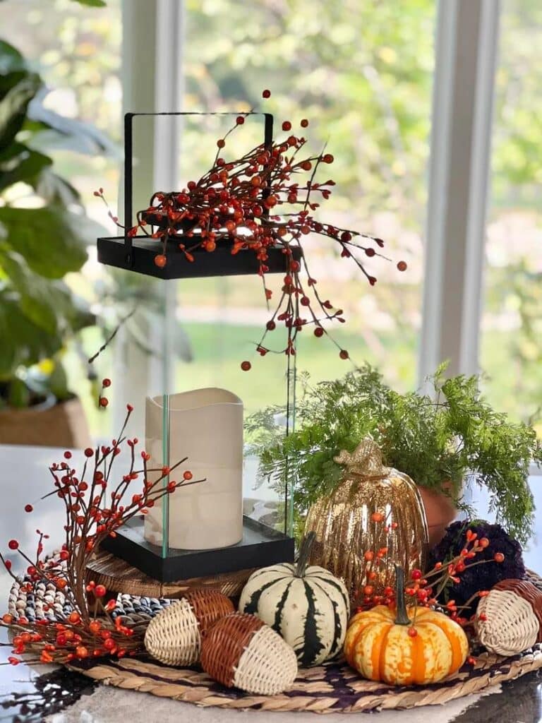 kitchen fall decor ideas: A glass lantern displayed on a woven mat with small pumpkins and rattan acorns.