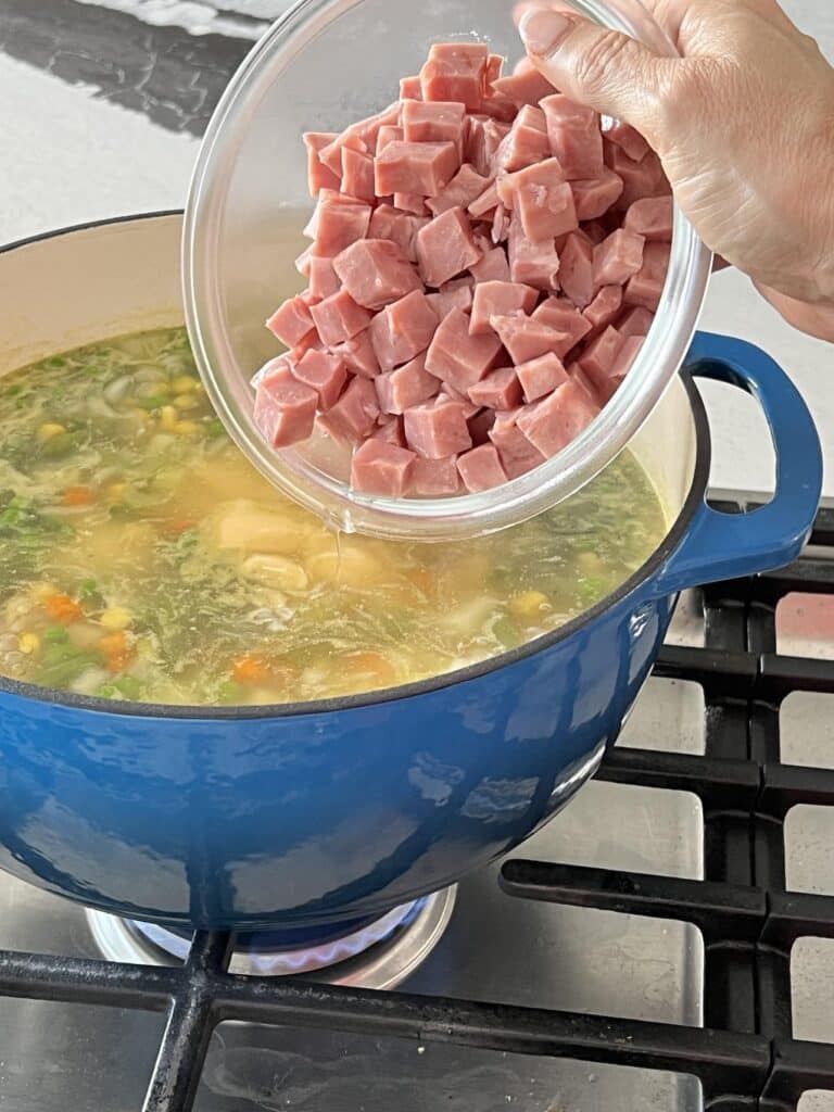 Adding cubed ham to a blue pot of brown jug soup.